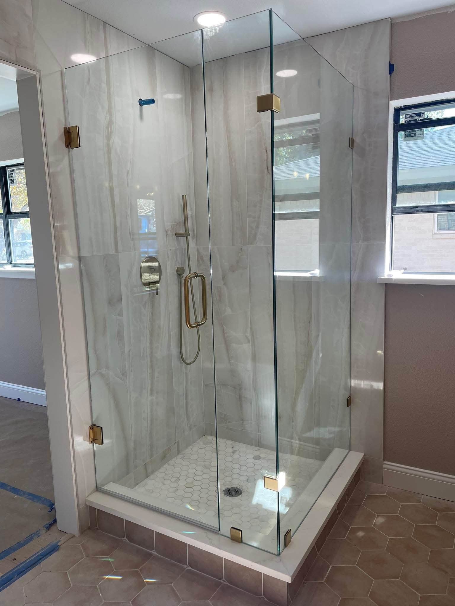 Modern glass shower enclosure featuring gold hardware, marble-patterned walls, and white hexagonal floor tiles.